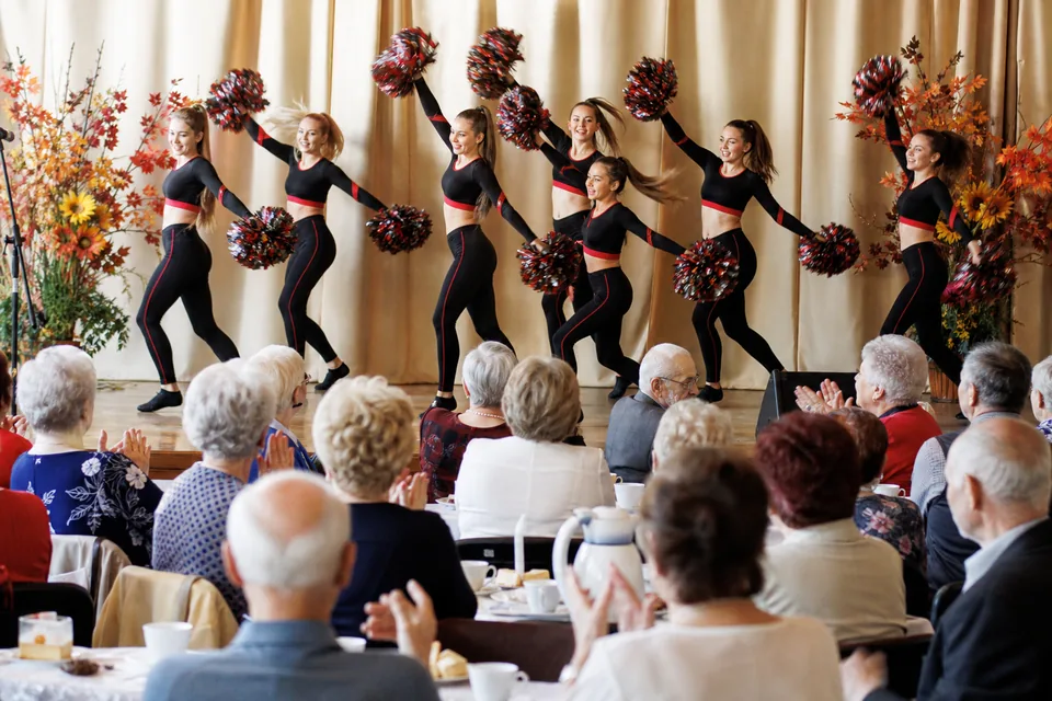 Seniorzy przy stołach w sali ośrodka kultury oglądają pokaz grupy tanecznej z pomponami